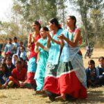 Vier ehemalige Kamalari führen vor dem Beginn eines Theaterstücks einen Tanz auf. Das Stück soll die Schüler auf die Probleme der Kamalari-Mädchen aufmerksam machen. Im Hintergrund sitzen weitere Mädchen und Jungen und sehen zu