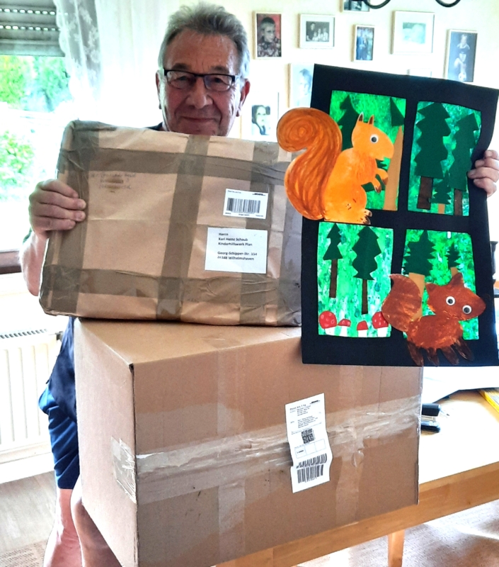 Ein älterer Mann mit Brille steht im Haus, lächelt und hält zwei große braune Pappverpackungen in der Hand, die auf einem Tisch gestapelt sind. In der anderen Hand hält er ein buntes Gemälde von Eichhörnchen und Bäumen, das in vier Tafeln mit Grün-, Orange- und Brauntönen unterteilt ist.