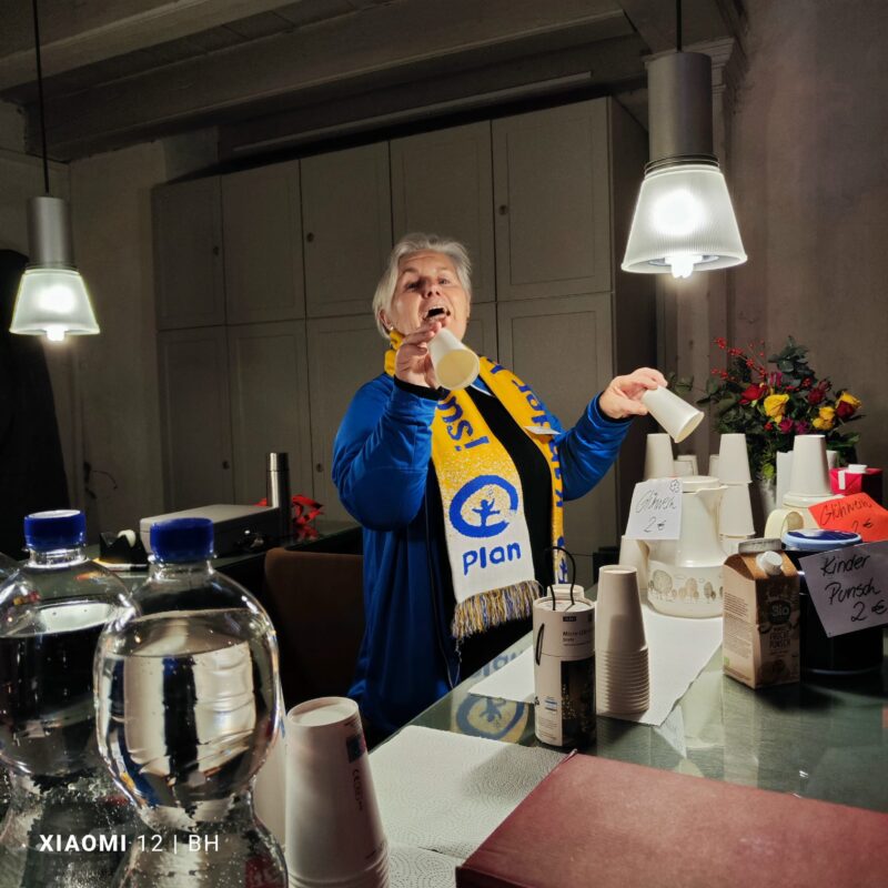 Eine ältere Frau in blauer Jacke und gelbem Schal steht hinter einem Tresen mit gestapelten Tassen, Getränken und Preisschildern. Sie hält einen weißen Becher in der Hand, während sie spricht, beleuchtet von Hängelampen. Wasserflaschen und Kartons sind zu sehen, im Hintergrund stehen Blumen.