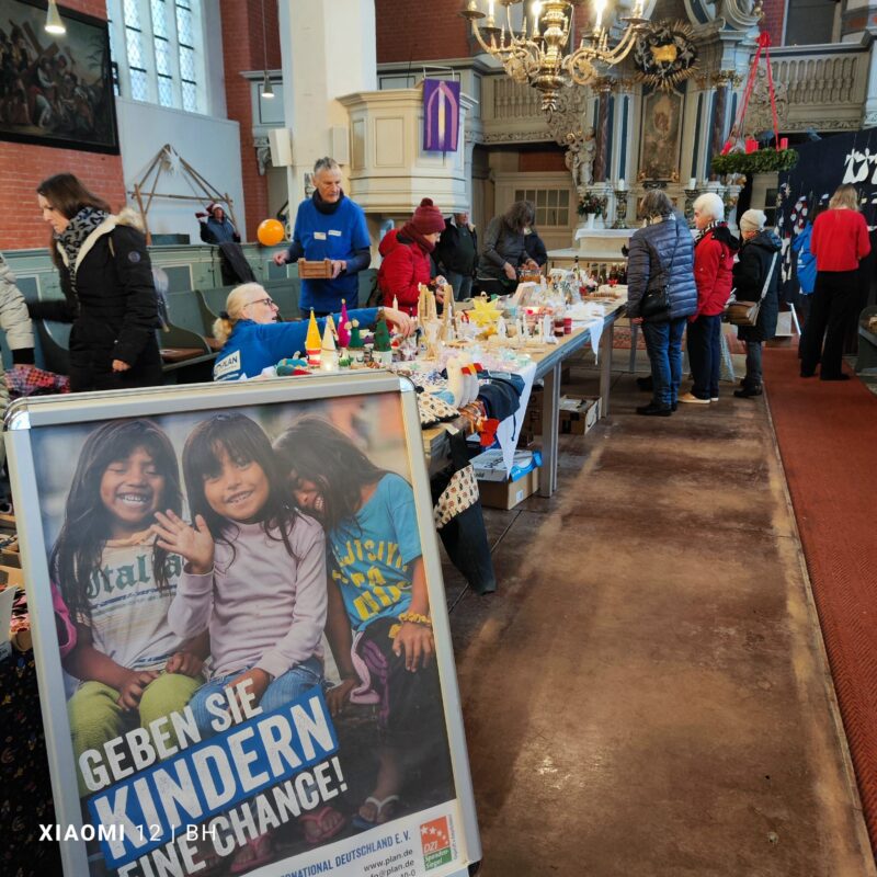 In einem Gemeindesaal findet ein belebter Wohltätigkeitsbasar statt, auf dem Tische mit Waren stehen und Menschen stöbern. Im Vordergrund zeigt ein Plakat drei lächelnde Kinder mit der Aufschrift "Geben Sie Kindern eine Chance!" (Gebt Kindern eine Chance!). Kronleuchter und Transparente schmücken den Raum.