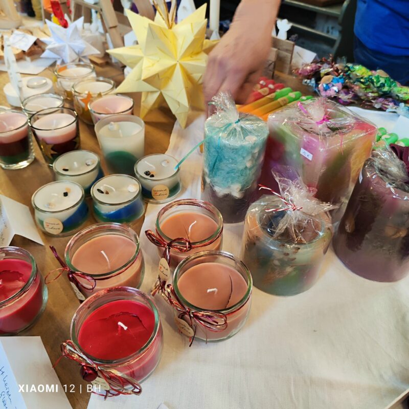 Ein Tisch mit bunten, handgefertigten Kerzen in Gläsern und verpackten Säulen steht auf einem hellen Tuch. Im Hintergrund sind Origami-Sterne, verpackte bunte Kerzen und eine Person zu sehen, die Gegenstände von Hand arrangiert.