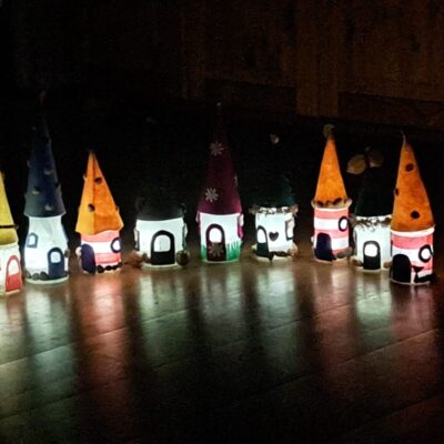 Eine Reihe kleiner, leuchtender, skurriler Häuser mit spitzen Dächern, die an Zwergen- oder Feenhäuser erinnern, steht auf einem Holzboden in einem dunklen Raum. Jedes Haus ist von innen beleuchtet und mit bunten Dächern und Fenstern verziert, die ein weiches, verspieltes Licht verbreiten.