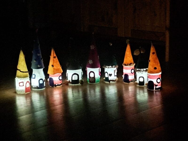 Eine Reihe kleiner, leuchtender, skurriler Häuser mit spitzen Dächern, die an Zwergen- oder Feenhäuser erinnern, steht auf einem Holzboden in einem dunklen Raum. Jedes Haus ist von innen beleuchtet und mit bunten Dächern und Fenstern verziert, die ein weiches, verspieltes Licht verbreiten.