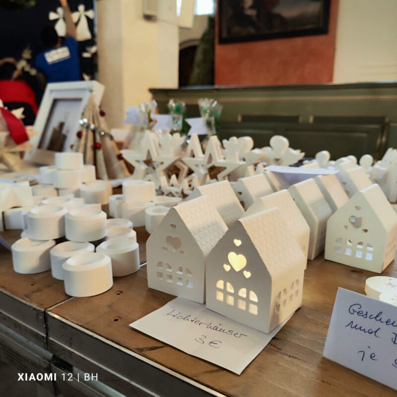 Auf einem Holztisch stehen kleine, weiße, dekorative Keramikhäuser mit herzförmigen Aussparungen. Einige Häuser sind von innen beleuchtet. Andere handgefertigte Produkte wie weiße LED-Teelichter und Holzsterne sind ebenfalls auf einem Markt in der Nähe ausgestellt.
