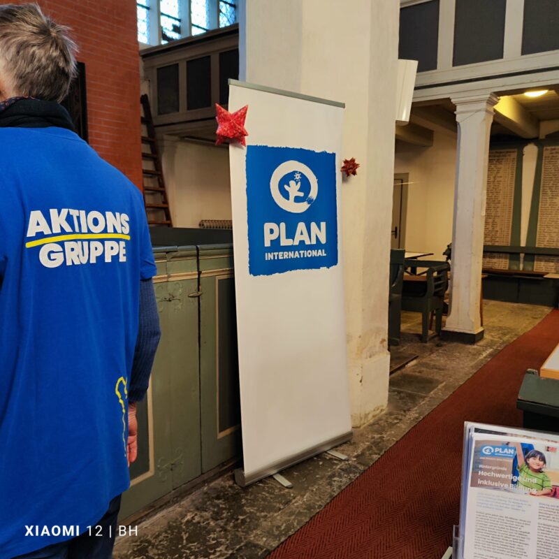 Eine Person in einem blauen AKTIONS-GRUPPE-Hemd steht neben einem PLAN International-Banner in einem kirchenähnlichen Gebäude. Die Szene enthält rote Sterndekorationen, einen Holzboden und Informationsbroschüren auf einem Tisch in der Nähe.
