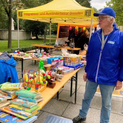 Ein älterer Mann in blauer Jacke und Mütze steht neben Tischen mit Kinderbüchern, Spielzeug und Spielen an einem Stand im Freien. Auf dem gelben Zelt hinter ihm steht Integrations- und Ausländerbeirat Dresden. Im Hintergrund sind Menschen und Bäume zu sehen.