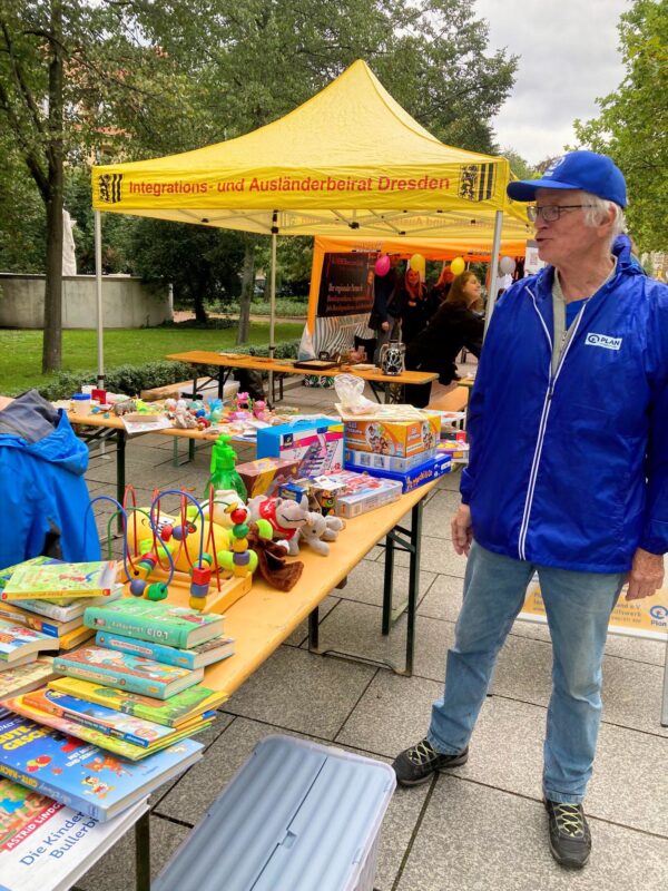 Ein älterer Mann in blauer Jacke und Mütze steht neben Tischen mit Kinderbüchern, Spielzeug und Spielen an einem Stand im Freien. Auf dem gelben Zelt hinter ihm steht Integrations- und Ausländerbeirat Dresden. Im Hintergrund sind Menschen und Bäume zu sehen.