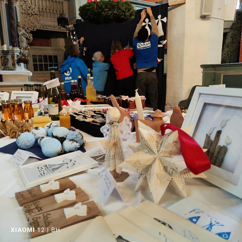 Auf einem festlich geschmückten Markttisch werden handgefertigtes Kunsthandwerk, Ornamente, gerahmte Kunstwerke, Getränke in Flaschen und Backwaren angeboten. Im Hintergrund hängen drei Personen in blauen Hemden Dekorationen vor einem dunklen Hintergrund mit einem Kranz darüber auf und bereiten sich auf eine Veranstaltung im Haus vor.
