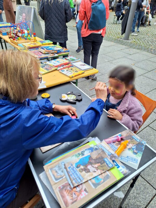 Ein Erwachsener mit blondem Haar und Brille, der eine blaue Jacke trägt, bemalt das Gesicht eines kleinen Kindes bei einer Veranstaltung im Freien. Das Kind sitzt an einem Tisch, der mit Tierzeitschriften bedeckt ist. Dahinter stehen weitere Tische mit bunten Büchern und Spielzeug, während die Menschen auf den Kopfsteinpflasterwegen spazieren gehen.