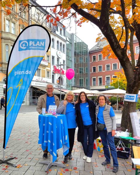 Vier lächelnde Menschen in blauen Hemden stehen an einem Infostand von Plan International im Freien, unter herbstlich gefärbten Blättern. Ihr Stand ist mit Broschüren und einer Markenfahne ausgestattet. Im Hintergrund sind historische Gebäude und Marktstände zu sehen.