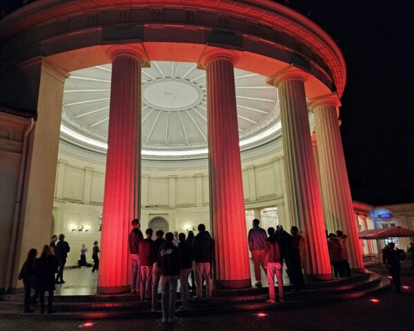 Eine Menschenmenge steht vor einem großen, runden neoklassizistischen Gebäude mit hohen Säulen, die nachts von roten Lichtern beleuchtet werden. Die Kuppel und die kunstvollen Details des Gebäudes sind sichtbar, während das Innere sanft beleuchtet ist. Die Szene ist lebhaft und deutet auf eine Veranstaltung oder ein Treffen hin.