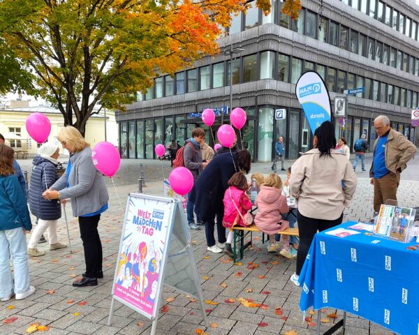 Menschen versammeln sich im Freien vor einem mit einem blauen Tuch bedeckten Tisch für eine Veranstaltung. Kinder halten rosa Luftballons, während Erwachsene sie beaufsichtigen und ihnen helfen. Ein Informationsschild und bunte Blätter an Bäumen schaffen eine lebendige Atmosphäre neben einem modernen Gebäude.