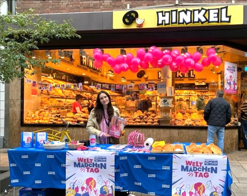 Eine Frau lächelt hinter einem blauen Tisch mit Flyern, Tassen und Dekorationen für den "Welt Mädchen Tag" vor einer Bäckerei namens Hinkel. Darüber schweben rosafarbene Luftballons, und im Schaufenster der Bäckerei sind Backwaren ausgestellt.