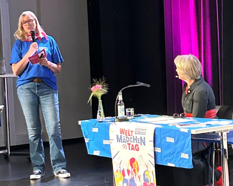 Eine Frau in blauem Hemd und Jeans spricht in ein Mikrofon, während sie neben einem Tisch steht. Eine andere Frau, die an dem Tisch sitzt, hört zu. Der Tisch ist mit einem blauen Tuch und einem bunten Schild mit der Aufschrift "Welt Mädchen Tag" bedeckt. Im Hintergrund ist eine rosa beleuchtete Wand zu sehen.