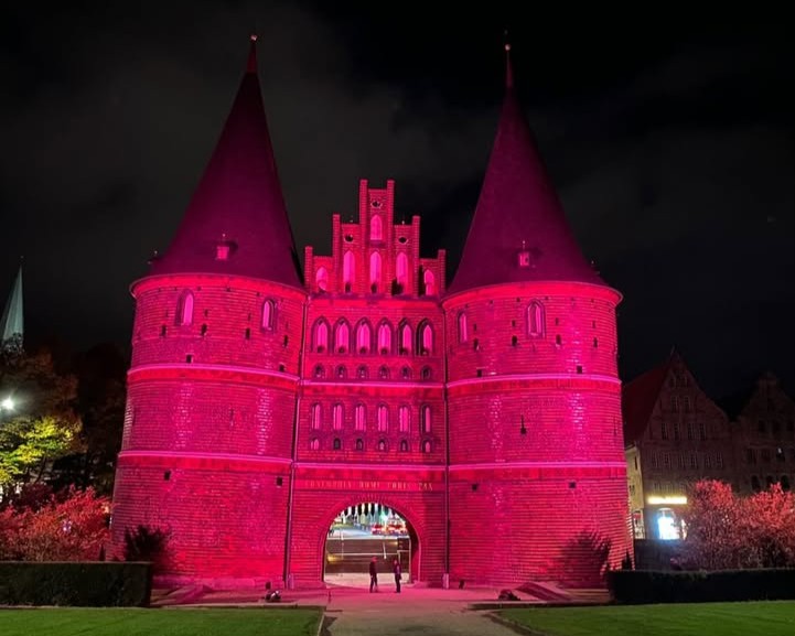Ein historisches Stadttor aus Backstein mit zwei hohen, spitzen Türmen wird nachts mit hellen rosa Lichtern dramatisch beleuchtet. Zwei kleine Figuren stehen unter dem Torbogen. Der Himmel ist dunkel, und die umliegenden Gebäude sind nur schwach zu erkennen.