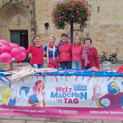 Fünf Frauen stehen lächelnd hinter einem mit rosa Luftballons und Informationsmaterial geschmückten Tisch vor einem historischen Steingebäude. Auf einem hellen Banner steht "Weltmädchentag" und wirbt für den Internationalen Tag des Mädchens am 11. Oktober.
