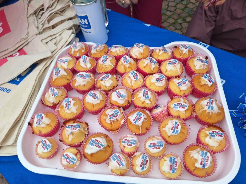 Ein Tablett mit Cupcakes mit weißem Zuckerguss, auf denen jeweils ein kreisförmiger essbarer Aufdruck mit der Aufschrift "Mein Name, Mein Tag" angebracht ist. Die Cupcakes sind in rosafarbenen und weißen Papierhüllen auf einem weißen Tablett auf einem blauen Tisch mit beigen Tragetaschen in der Nähe angeordnet.