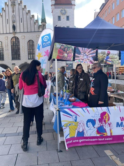 Menschen versammeln sich an einem Veranstaltungsstand im Freien, der mit bunten Bannern für Gleichberechtigung wirbt. Zwei Frauen stehen hinter dem Tisch mit Broschüren und blauen Luftballons, während ein Besucher in einer rosa Jacke zuhört. Im Hintergrund sind historische Gebäude und eine Menschenmenge zu sehen.