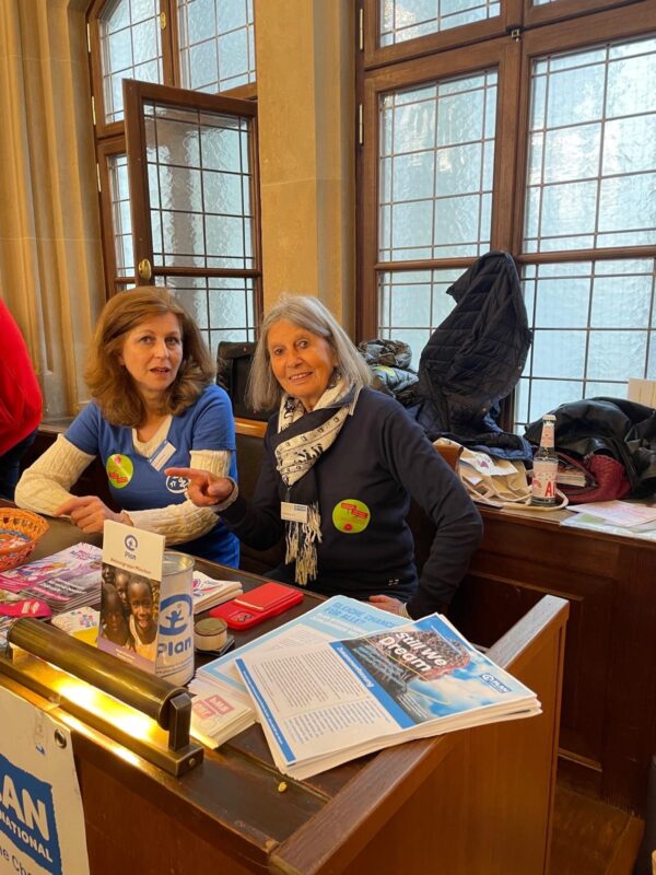 Zwei Frauen sitzen an einem Holztisch mit Flugblättern, Zeitschriften und einer Spendenbox. Beide tragen blaue Hemden mit bunten Knöpfen und lächeln in die Kamera. Hinter ihnen befinden sich hohe Fenster mit Milchglas und Mäntel, die auf Stühlen hängen.