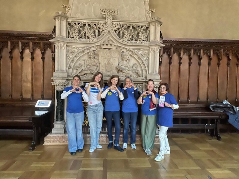 Sieben Frauen stehen Seite an Seite vor einem verzierten Steinaltar, tragen blaue Hemden und lächeln. Einige bilden mit ihren Händen Herzformen. Der Raum ist holzgetäfelt, hat einen polierten Holzboden und eine Bank links von der Gruppe.