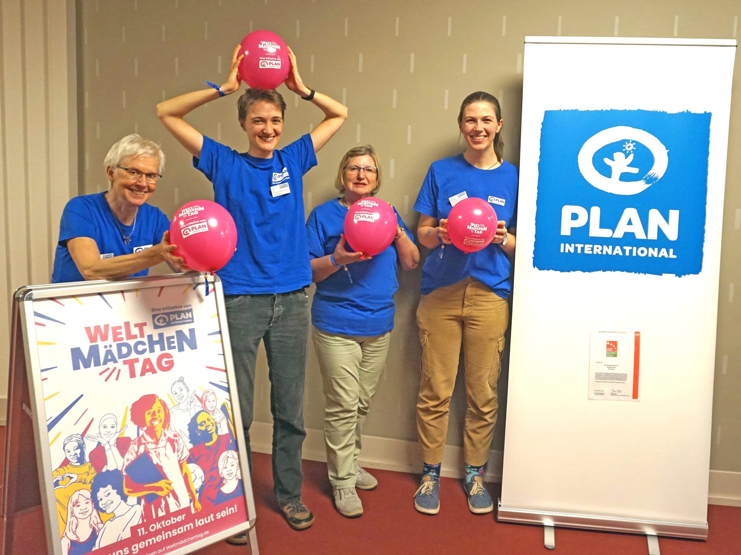 Vier Personen in blauen Plan International-Hemden stehen in einem Innenraum, lächeln und halten rosa Luftballons. Zwei Transparente in der Nähe zeigen das Logo von Plan International und Informationen zum Welt Mädchentag. Die Stimmung ist fröhlich und feierlich.