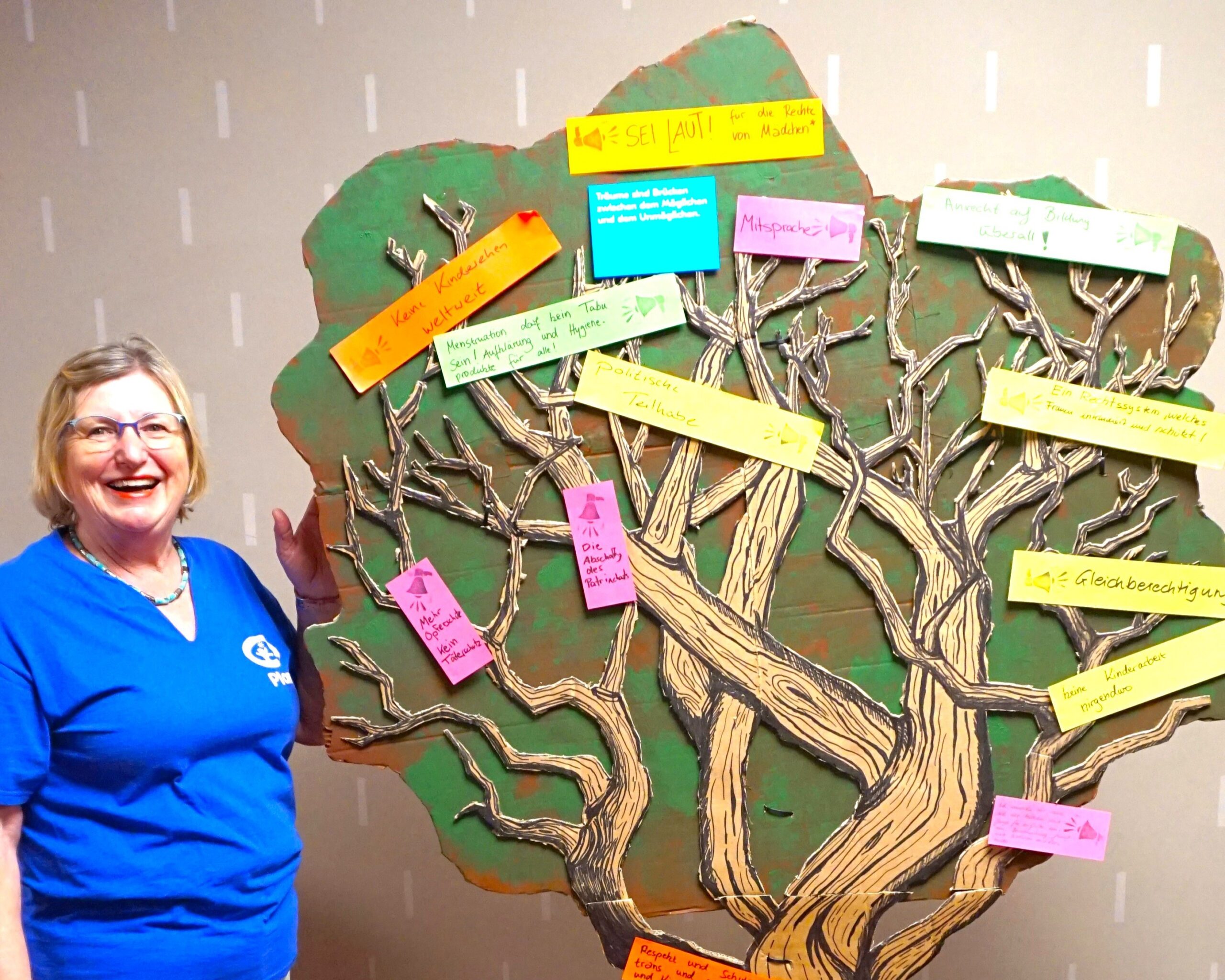 Eine lächelnde Frau in einem blauen Hemd steht neben einem gemalten Baum, der an einer Wand hängt. Der Baum hat viele Äste, an denen bunte Papierzettel mit handgeschriebenem Text befestigt sind. Der Hintergrund ist einfarbig mit vertikalen weißen Strichen.