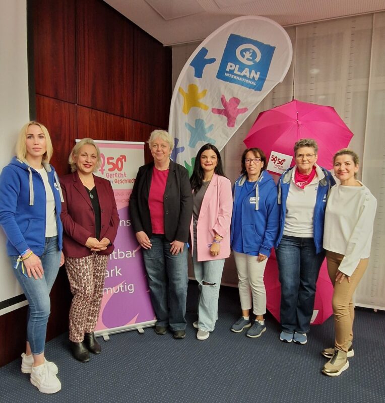 Sieben Frauen stehen drinnen in Pose und lächeln vor Plan International und 50/50-Bannern. Sie tragen eine Mischung aus Freizeit- und Geschäftskleidung, einige in blauen Kapuzenpullis, andere in Blazern. Im Hintergrund sind ein rosa Regenschirm und eine Markenflagge zu sehen.
