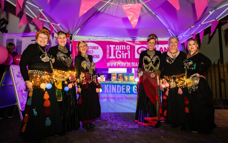 Sechs Frauen in bunten, traditionellen Bauchtanzkostümen mit dekorativen Accessoires und Lichterketten posieren und lächeln unter einem Zelt. Der Hintergrund ist in Rosa und Lila gehalten und zeigt Banner, die für Plan International und die Kampagne Because I am a Girl werben.