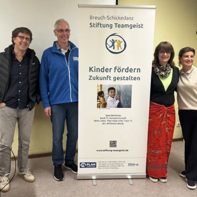 Vier Erwachsene stehen lächelnd neben einem Transparent der Stiftung Teamgeist. Das Banner trägt die Aufschrift "Kinder fördern, Zukunft gestalten" und zeigt ein Foto eines glücklichen Kindes. Die Gruppe steht in einem ungezwungenen Innenraum und zeigt Einigkeit und Unterstützung für die Organisation.