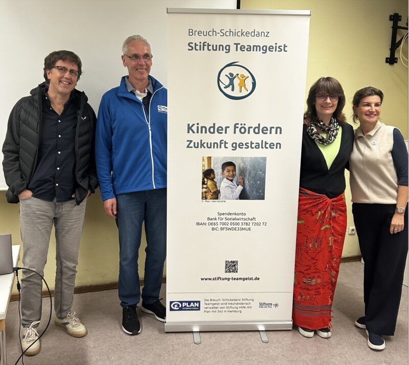 Vier Erwachsene stehen lächelnd neben einem Transparent der Stiftung Teamgeist. Das Banner trägt die Aufschrift "Kinder fördern, Zukunft gestalten" und zeigt ein Foto eines glücklichen Kindes. Die Gruppe steht in einem ungezwungenen Innenraum und zeigt Einigkeit und Unterstützung für die Organisation.