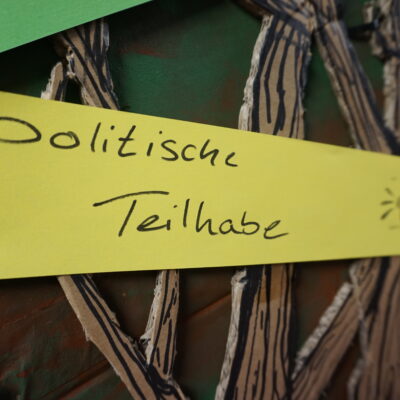 Ein gelber Papierstreifen mit der handschriftlichen Aufschrift "Politische Teilhabe" und einem kleinen Megaphon-Symbol ist auf einen Hintergrund gepinnt, der mit braunen, baumzweigartigen Pappausschnitten verziert ist. Das Foto ist stark beschnitten.