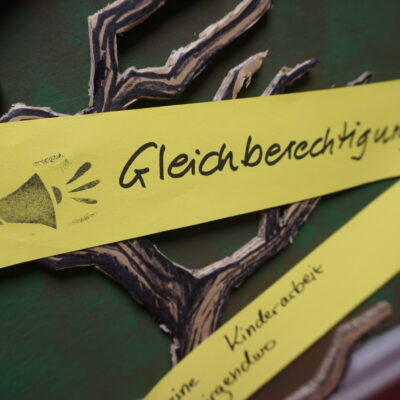 Ein gelber Papierstreifen mit einem gezeichneten Megaphon und dem deutschen Wort "Gleichberechtigung" ist an ein Display in Form eines Astes gepinnt. Darunter befindet sich ein weiterer gelber Streifen mit teilweise sichtbarem Text.