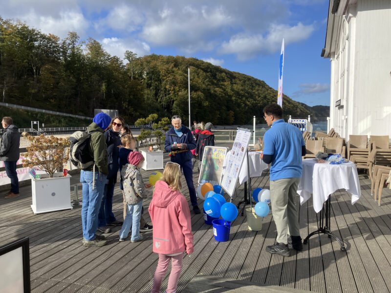 Eine Gruppe von Erwachsenen und Kindern versammelt sich im Freien auf einem Holzdeck in der Nähe eines weißen Gebäudes. Einige sehen sich Informationsdisplays auf Tischen mit blauen und gelben Luftballons an. Im Hintergrund sind Bäume und Hügel zu sehen, der Himmel ist teilweise bewölkt.