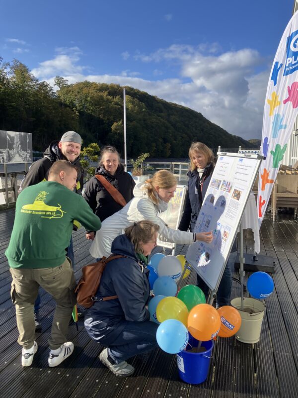 Sechs Erwachsene versammeln sich auf einer Holzterrasse im Freien, lächeln und interagieren mit einer Anzeigetafel. Bunte Luftballons und Informationsposter sind zu sehen. Eine weiße Werbefahne und Eimer mit Luftballons stehen in der Nähe. Grüne Bäume und ein blauer Himmel bilden den malerischen Hintergrund.