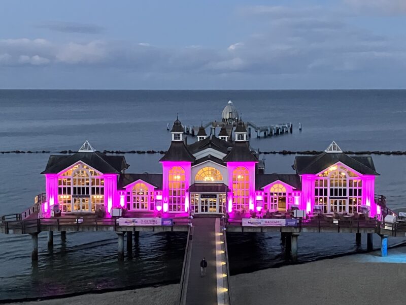 Ein großes Pier-Gebäude mit verzierten Dächern und vielen Fenstern ist mit leuchtend rosa Lichtern beleuchtet und steht in der Abenddämmerung über dem Wasser. Ein Holzsteg führt zum Eingang, und in der Ferne erstreckt sich das ruhige Meer unter einem bewölkten Himmel.