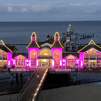 Ein großes Piergebäude über dem Meer wird in der Abenddämmerung mit hellen violetten und gelben Lichtern beleuchtet. Das Gebäude mit hohen Giebeldächern und großen Fenstern liegt über dem Wasser und ist durch eine Promenade mit dem Land verbunden. Im Hintergrund sind das ruhige Meer und der bewölkte Himmel zu sehen.