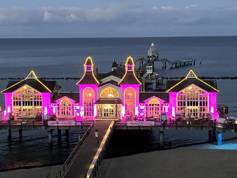 Ein großes Piergebäude über dem Meer wird in der Abenddämmerung mit hellen violetten und gelben Lichtern beleuchtet. Das Gebäude mit hohen Giebeldächern und großen Fenstern liegt über dem Wasser und ist durch eine Promenade mit dem Land verbunden. Im Hintergrund sind das ruhige Meer und der bewölkte Himmel zu sehen.