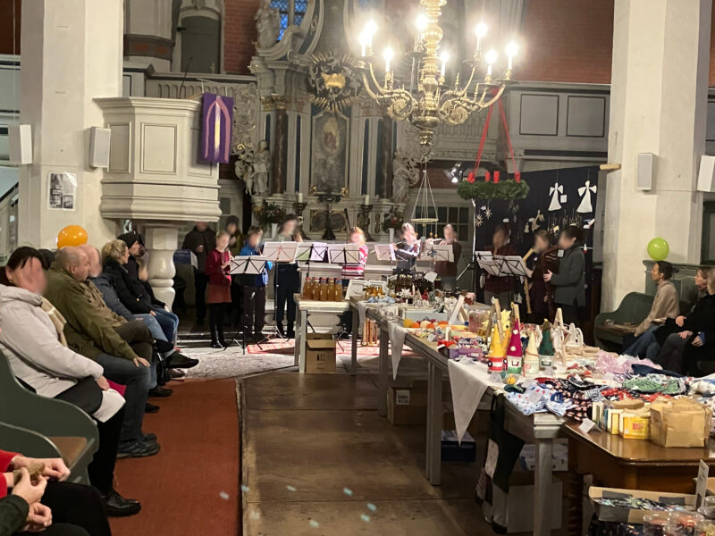 Eine Gruppe von Musikern spielt in einer geschmückten Kirche vor einem Publikum, das auf den Kirchenbänken sitzt. Auf den Tischen sind Kunsthandwerk, Geschenke und Einmachgläser ausgestellt. Festliche Dekorationen, Luftballons und ein großer Kronleuchter schaffen eine warme, lebendige Atmosphäre.