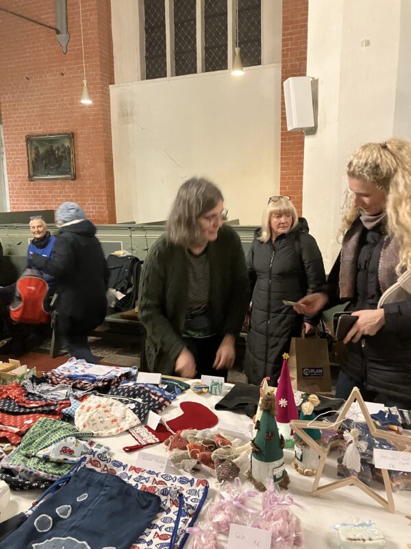Mehrere Frauen stöbern auf einem festlichen Kunsthandwerkermarkt in einer Halle, die mit handgefertigten Artikeln wie Stoffbeuteln, Puppen, Ornamenten und Dekorationen gedeckt ist. Im Hintergrund sind Backsteinwände, hohe Fenster und sitzende Besucher zu sehen, die eine gemütliche, lebhafte Szene schaffen.