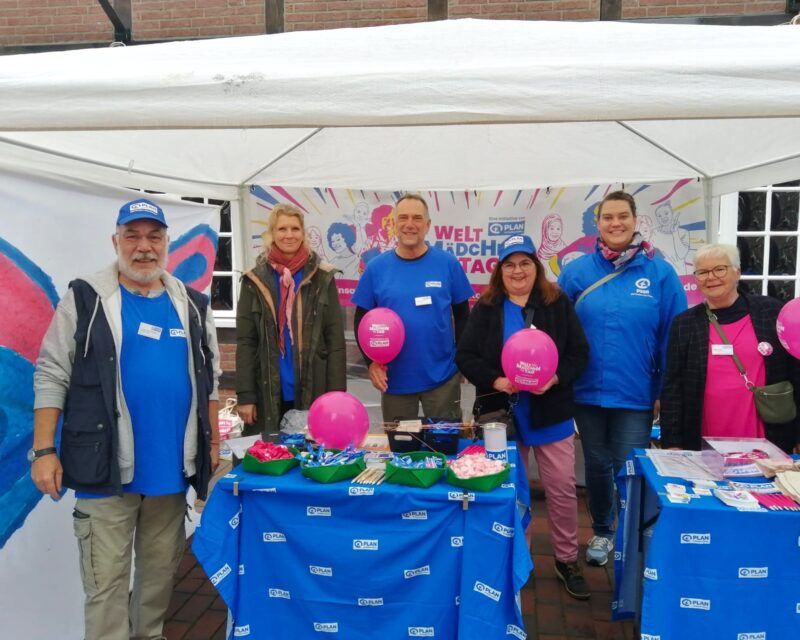 Sechs Personen stehen unter einem weißen Zelt bei einer Freiluftveranstaltung und lächeln in die Kamera. Sie tragen blaue und rosa Kleidung mit Logos und halten rosa Luftballons. Auf dem Tisch davor liegen Flyer, Süßigkeiten und Luftballons. Hinter ihnen hängt ein buntes Transparent.