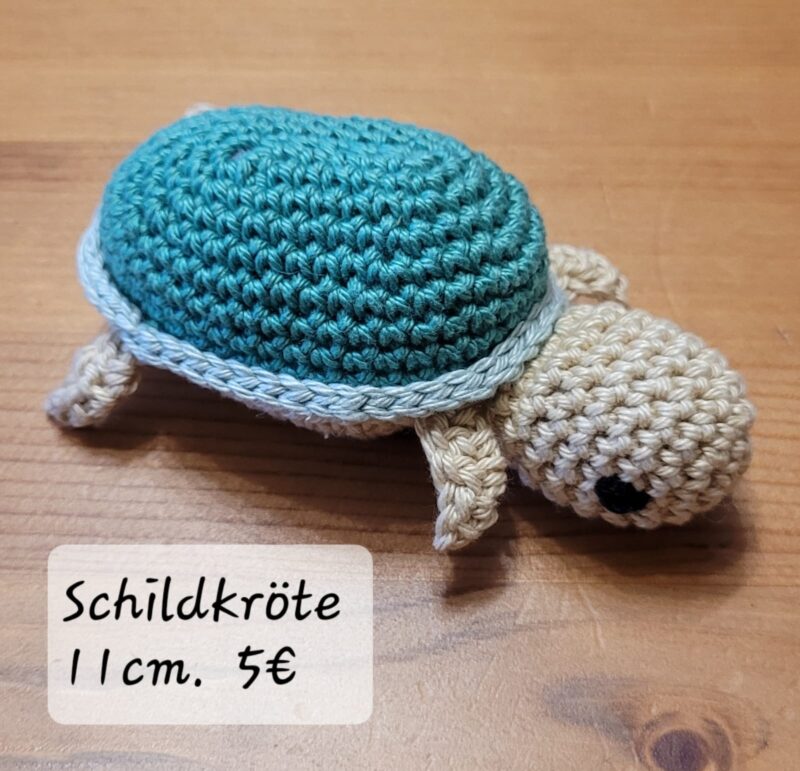 Eine kleine gehäkelte Schildkröte mit grünem Panzer und beigem Kopf, Beinen und Schwanz liegt auf einer Holzfläche. Auf einem Etikett in der Ecke steht "Schildkröte 11 cm. 5€" in handschriftlicher Schrift.