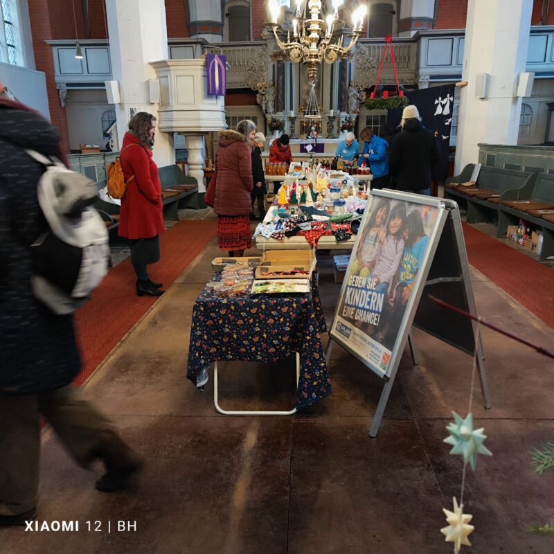 Ein Kirchenraum mit Menschen, die auf Tischen verschiedene handgefertigte Waren zum Verkauf anbieten. Im Vordergrund ist ein großes Schild in deutscher Sprache zu sehen, darüber hängt ein Kronleuchter. Im Hintergrund sind Bänke, eine Kanzel und dekorative Elemente zu sehen.