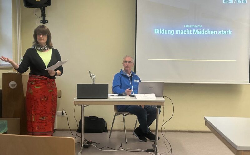 Eine Frau steht sprechend, mit Papieren in der Hand, neben einem sitzenden Mann mit einem Laptop. Hinter ihnen zeigt ein Projektor eine Präsentationsfolie auf Deutsch: Bildung macht Mädchen stark. Der Raum ist mit hellen Wänden und einfachen Möbeln ausgestattet, und die Einrichtung wirkt förmlich.