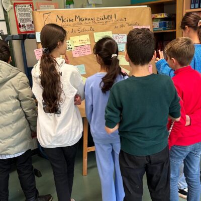 Sechs Kinder stehen mit dem Rücken zur Kamera und schauen auf eine braune Plakatwand mit handschriftlichen Notizen und Zeichnungen in einem Klassenzimmer. Regale, Papiere und bunte Poster schmücken den Raum um sie herum.