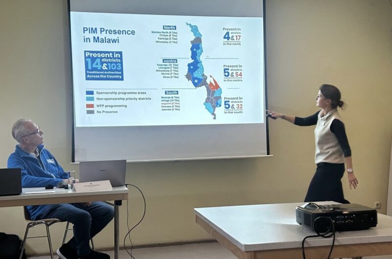 Eine Frau steht und zeigt auf eine projizierte Folie mit einer Karte und Statistiken über die Präsenz von PIM in Malawi, während ein Mann an einem Tisch mit einem Laptop sitzt. Die Folie zeigt Regionen, Zahlen und Text in Blau, Rot und Schwarz. Im Vordergrund befindet sich ein Projektor.