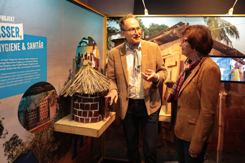 Zwei Personen in braunen Jacken und Schals stehen in einem Innenraum und unterhalten sich neben einer kleinen Modellhütte mit Strohdach. Hinter ihnen sind Ausstellungstafeln und Fotos über Wasser, Hygiene und sanitäre Einrichtungen in deutscher Sprache zu sehen. Die warme Beleuchtung unterstreicht die lehrreiche Umgebung.