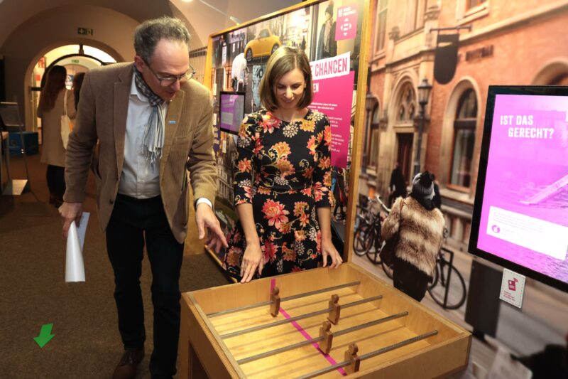 Ein Mann in einer braunen Jacke und einem Schal und eine Frau in einem geblümten Kleid stehen in einem Raum und schauen auf ein hölzernes Tischfußballspiel. An den Wänden hinter ihnen hängen bunte Poster und Fotos von Stadtszenen. Die Atmosphäre deutet auf eine Ausstellung oder Veranstaltung hin.