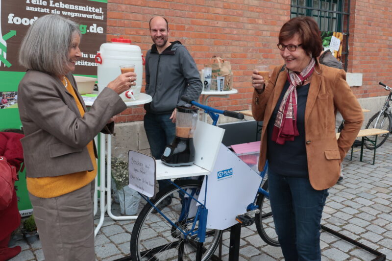 Zwei lächelnde Frauen stehen an einem mit dem Fahrrad betriebenen Smoothie-Mixer und stoßen mit Plastikbechern an. Ein Mann in einer grauen Jacke schaut lächelnd zu. Hinter ihnen stehen eine Backsteinmauer, ein grünes Foodsharing-Schild und ein Tisch mit Vorräten. Die Veranstaltung scheint festlich und im Freien zu sein.