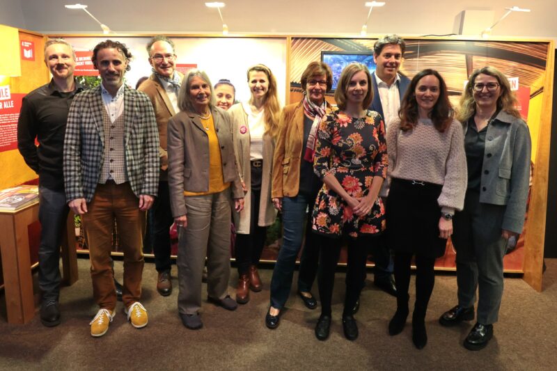 Eine Gruppe von elf Erwachsenen, Männer und Frauen, steht in zwei Reihen in einem Haus und lächelt in die Kamera. Sie sind leger gekleidet, und der Hintergrund besteht aus warmen Holzwänden, Plakaten und Oberlichtern, die der Szene eine freundliche, einladende Atmosphäre verleihen.