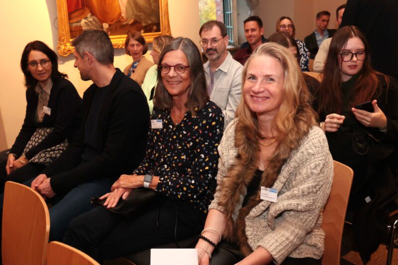 Eine Gruppe Erwachsener sitzt in einem gut beleuchteten Raum dicht beieinander, schaut nach vorne und lächelt. Einige halten Papiere oder Telefone in der Hand. Im Hintergrund sind ein goldgerahmtes Gemälde und Fenster zu sehen, was auf eine formelle oder berufliche Veranstaltung wie ein Seminar oder eine Präsentation hindeutet.